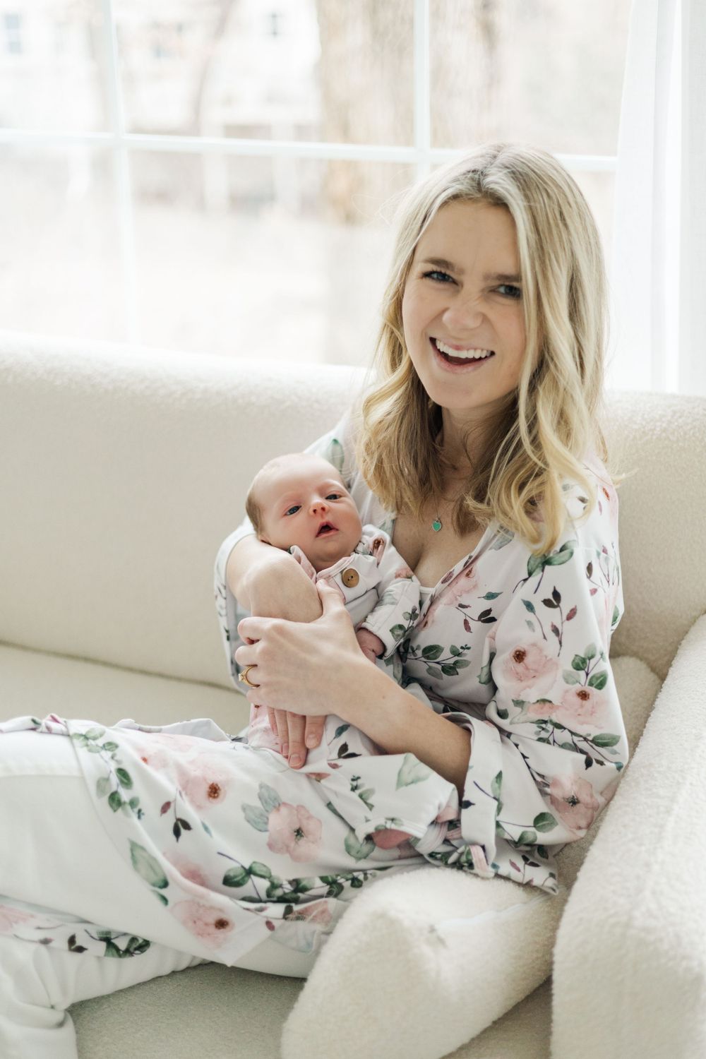 Parent and newborn in matching floral outfits relaxing on white couch in bright room.