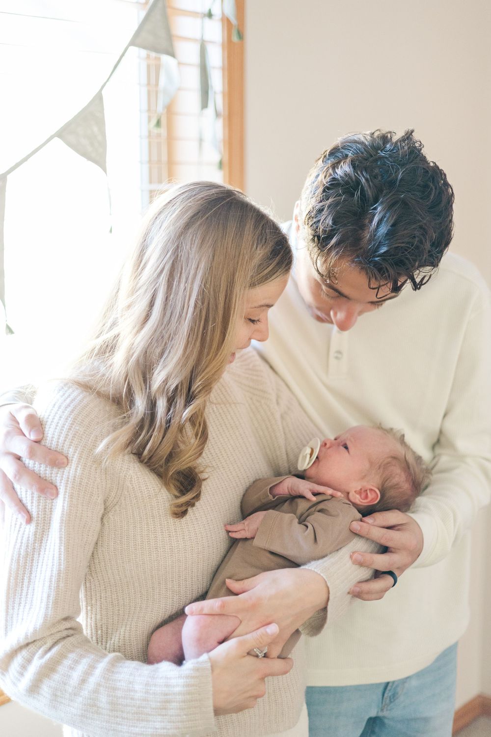 Parents in cozy sweaters tenderly holding their newborn baby near a bright window.