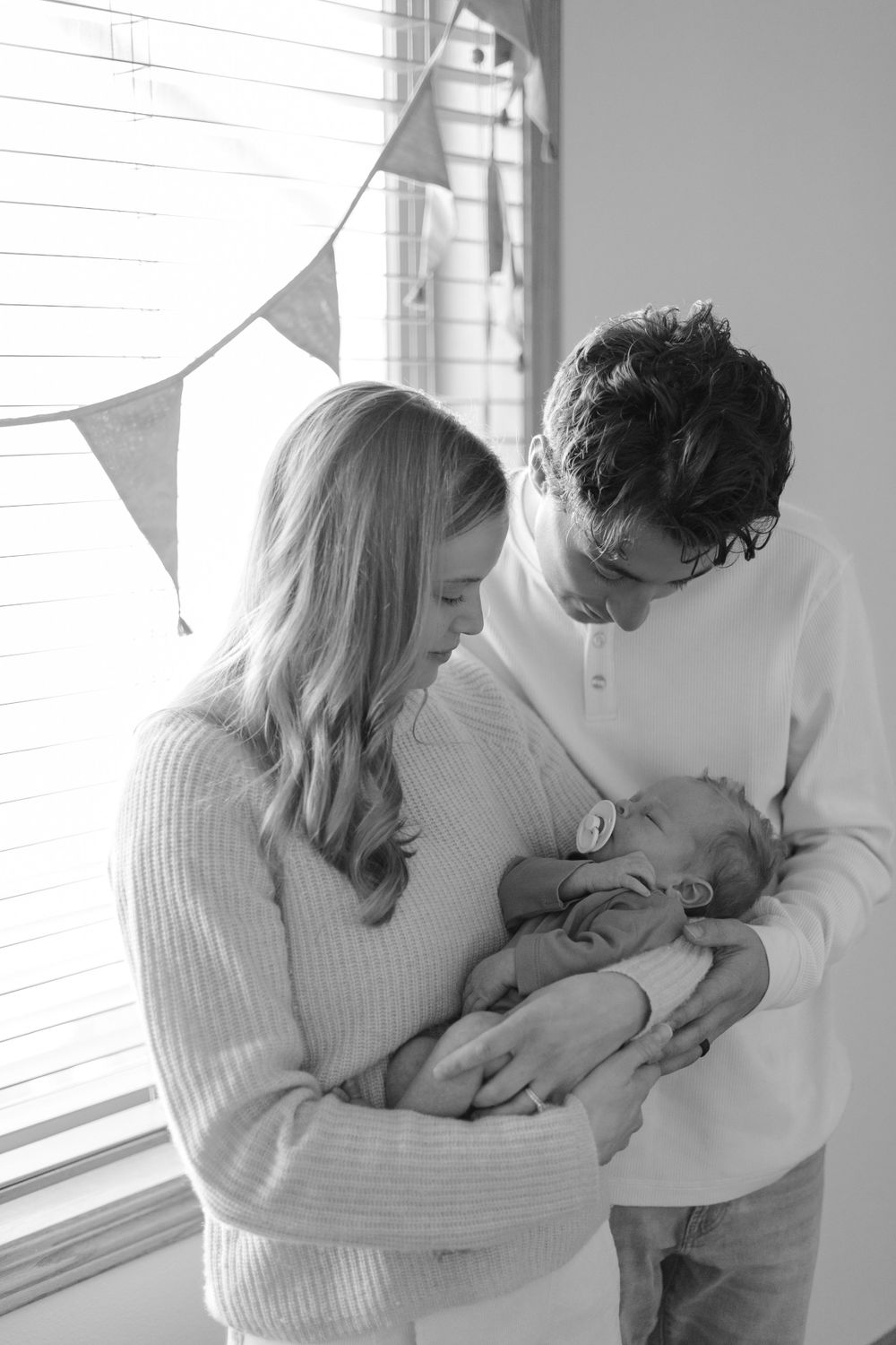 A tender black and white portrait of parents cradling their newborn by a window.