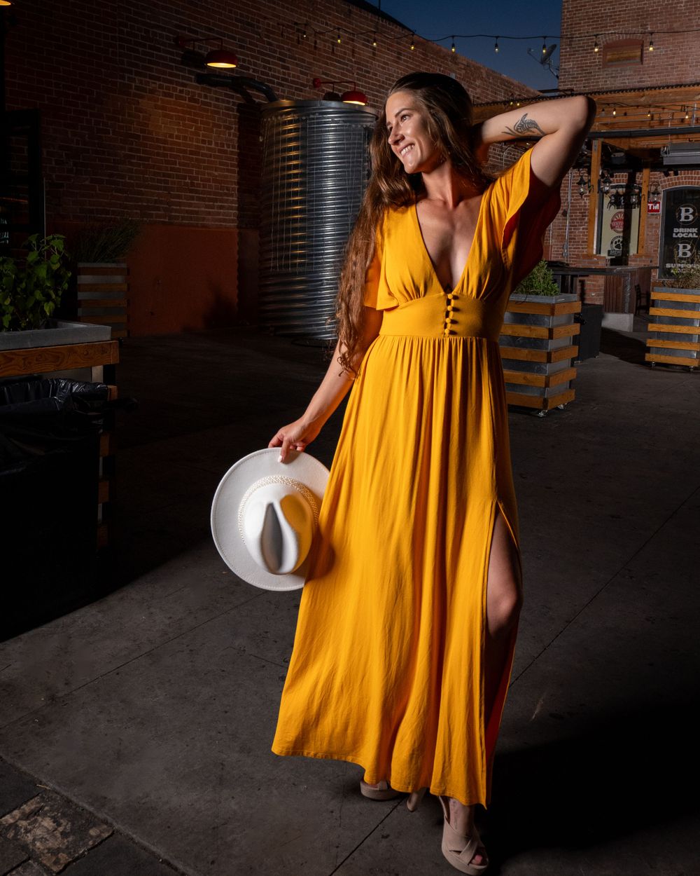Yellow maxi dress with thigh slit and white hat captured in industrial brick setting at sunset.
