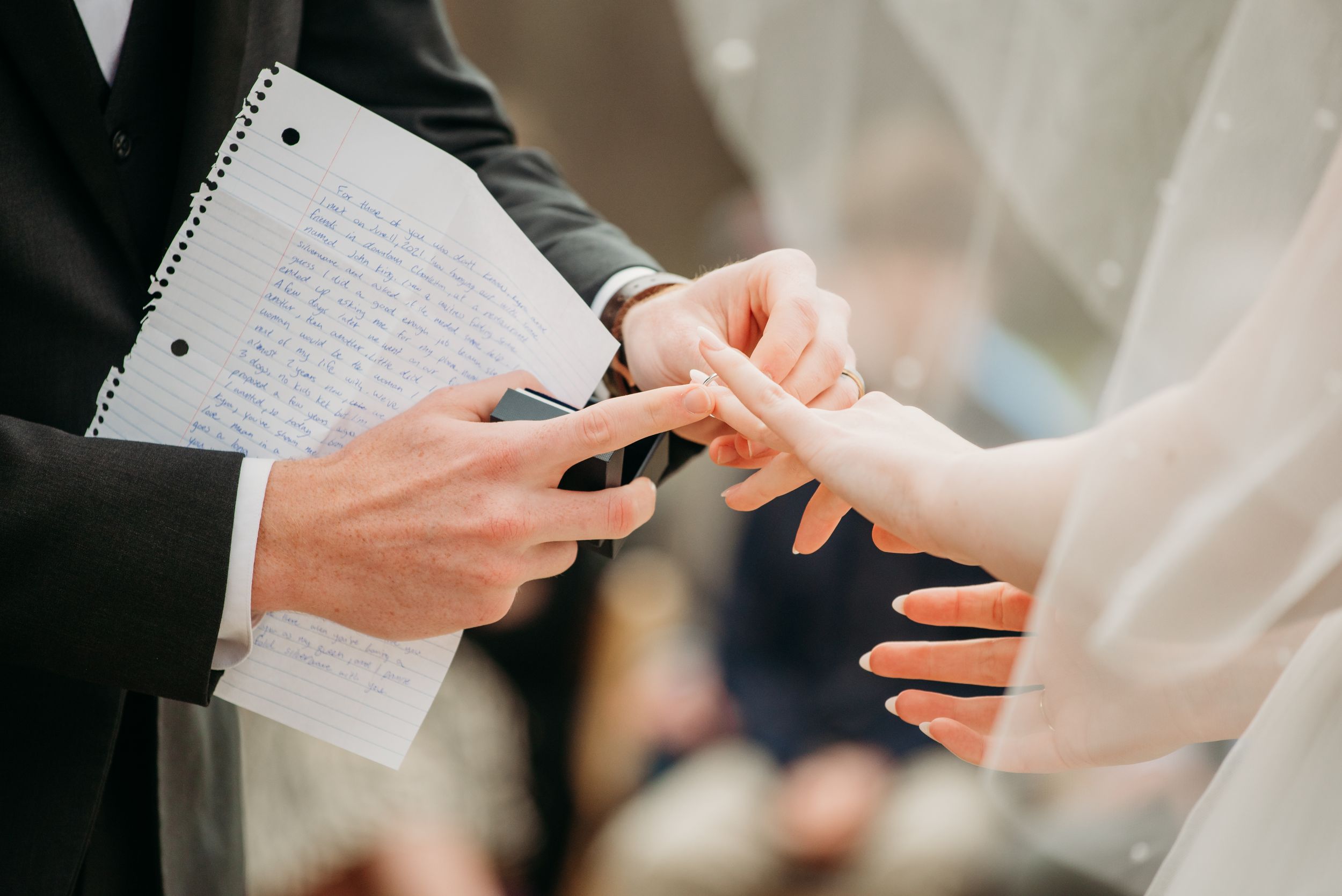 Close-up of hands exchanging rings during wedding ceremony with vows visible on notepad.