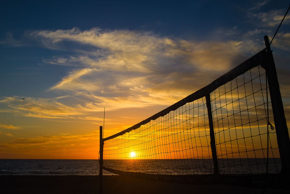 A beach volleyball net silhouetted against a vibrant orange sunset over the ocean creates a dramatic summer scene.