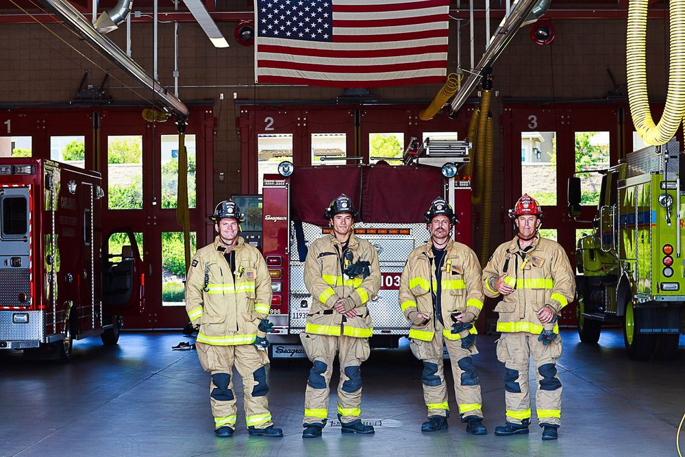 Firefighters in yellow reflective gear stand in formation at a fire station with American flags hanging overhead.