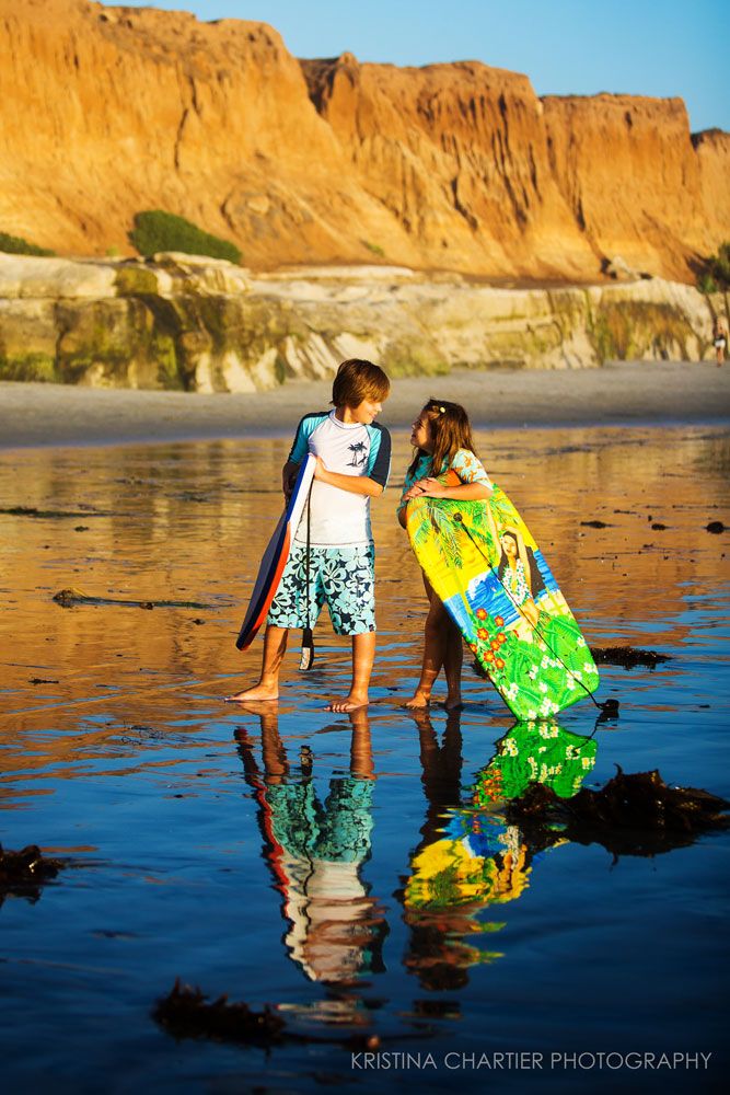Two young surfers walking on wet sand with colorful bodyboards at sunset.