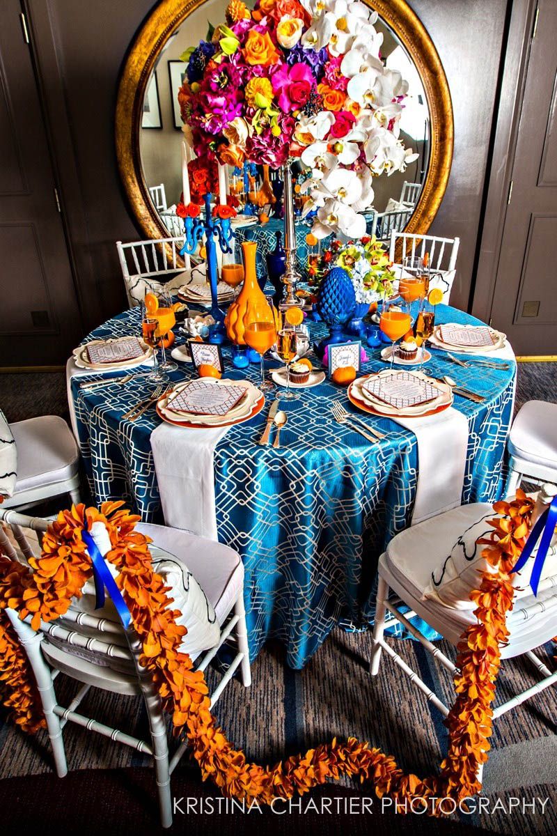 Elegant table setting with blue tablecloth, white chairs, and vibrant orange and purple floral centerpiece.