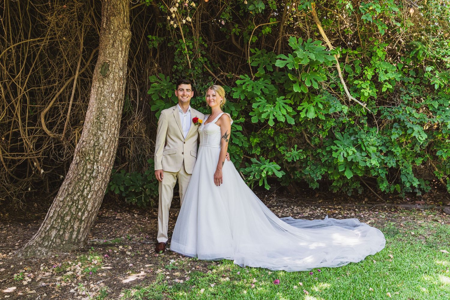 A couple in wedding attire poses together in a lush garden setting with trees and greenery.
