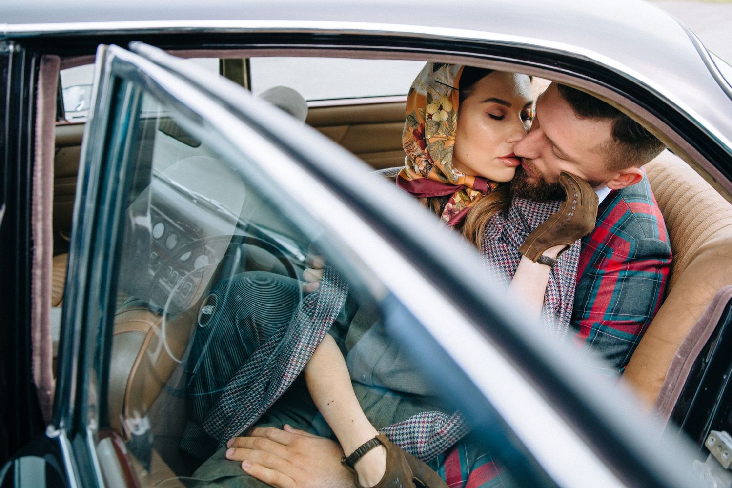 A romantic couple shares a tender kiss inside a vintage car parked on city street at dusk.
