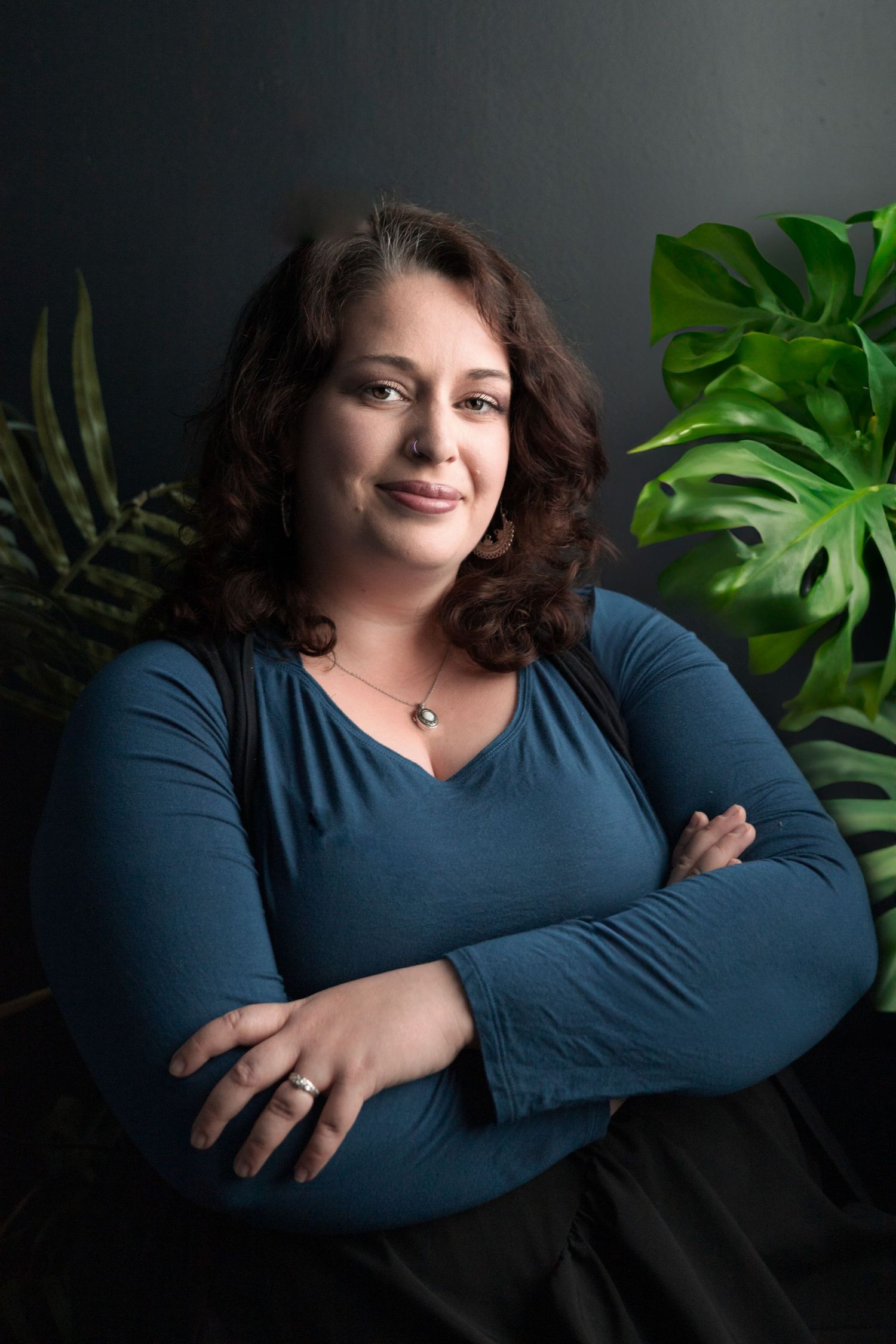 Portrait of a person in blue top with crossed arms posed against tropical plants and dark background.