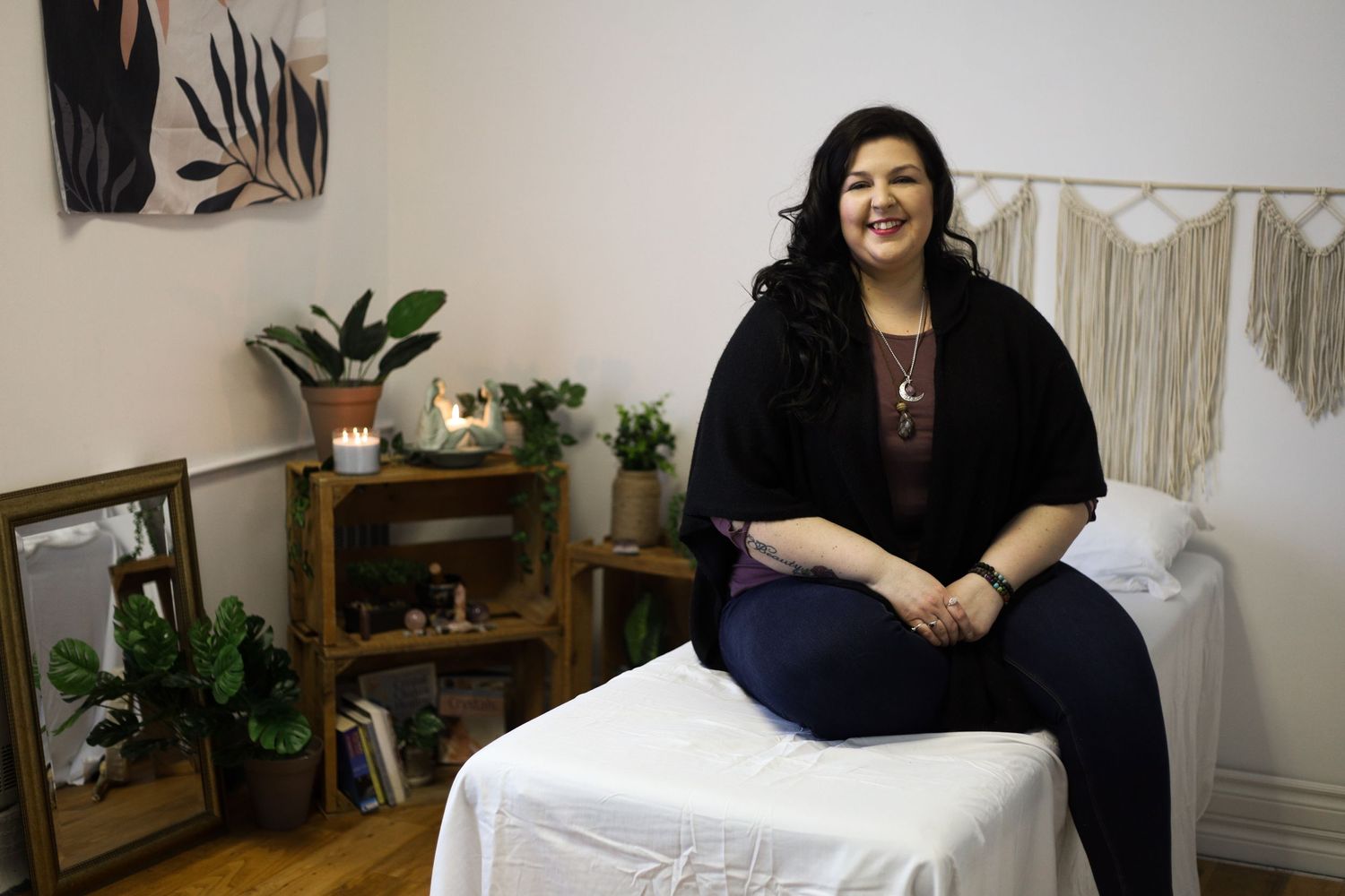 Lifestyle portrait of a person in black outfit seated in massage or treatment room with boho decor.