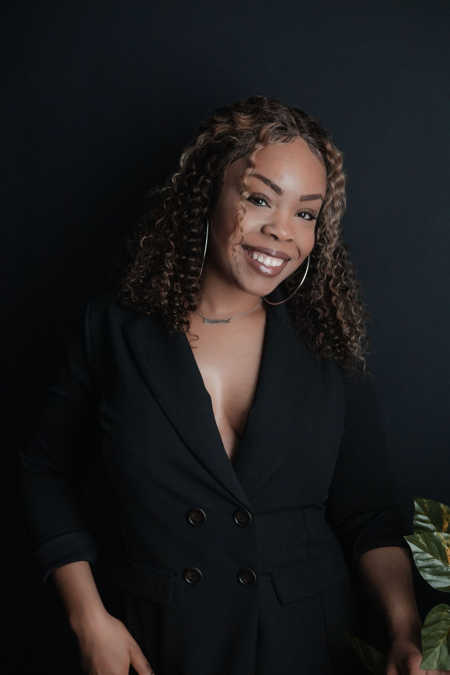 Professional portrait of a person in black blazer with curly hair against dark background with plant accent.