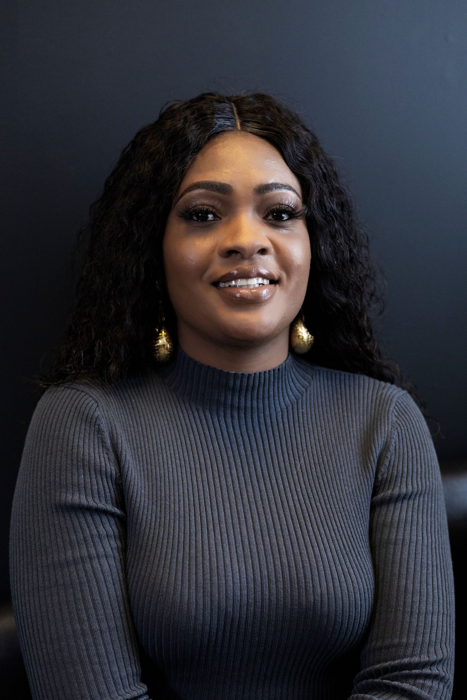 Studio portrait of a person in gray sweater wearing gold earrings with natural curly hair against dark background.