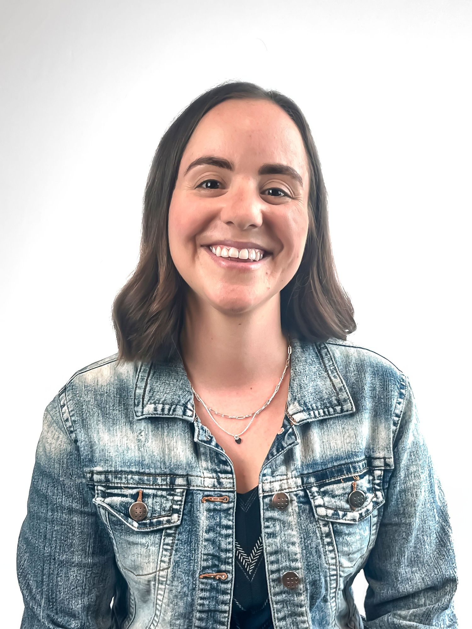 Portrait of someone wearing a light denim jacket and necklace against a white background while smiling warmly.