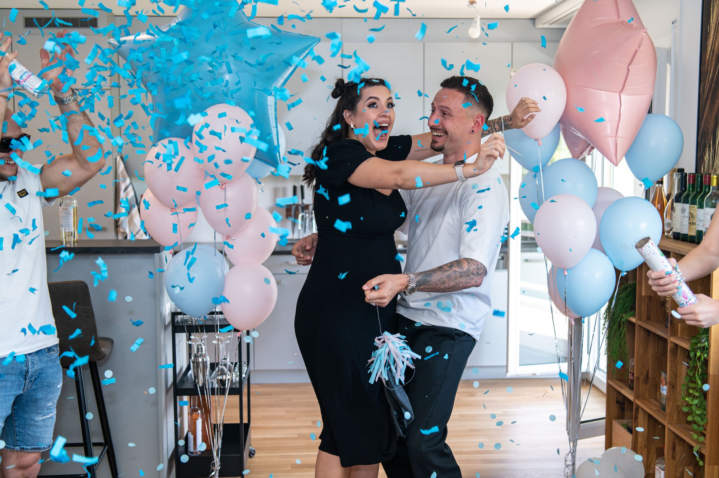 Joyful gender reveal moment with blue confetti and balloons in modern kitchen setting.