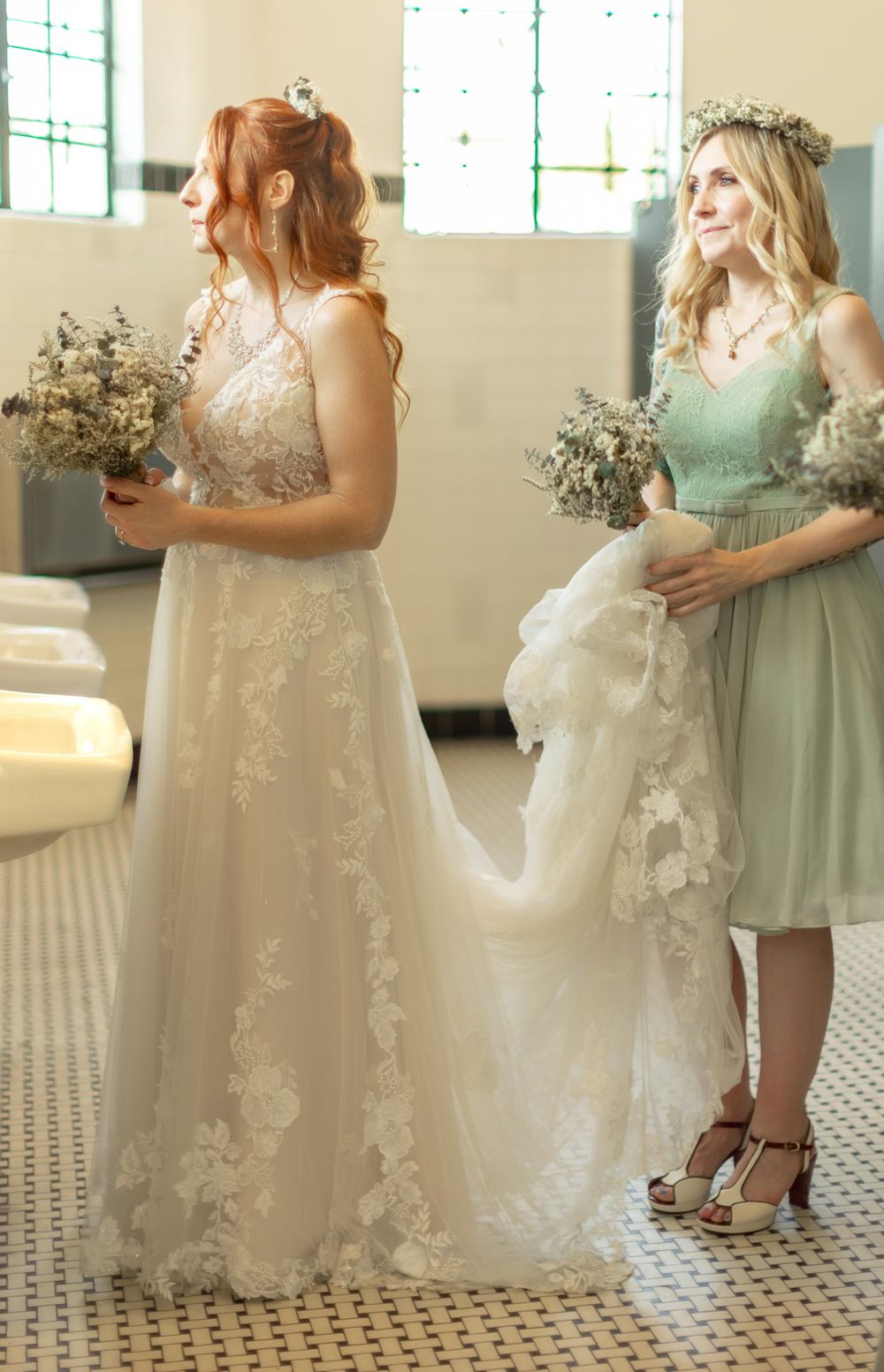 Elegant bridal scene with floral lace wedding gown and mint green bridesmaid dress holding bouquets in sunlit room.