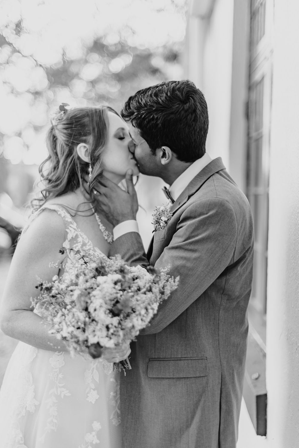 A black and white wedding photo captures a romantic kiss moment with the bride holding a delicate floral bouquet.