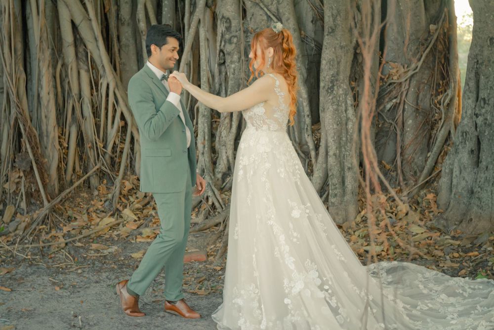 A couple in wedding attire dance together among banyan trees at sunset.
