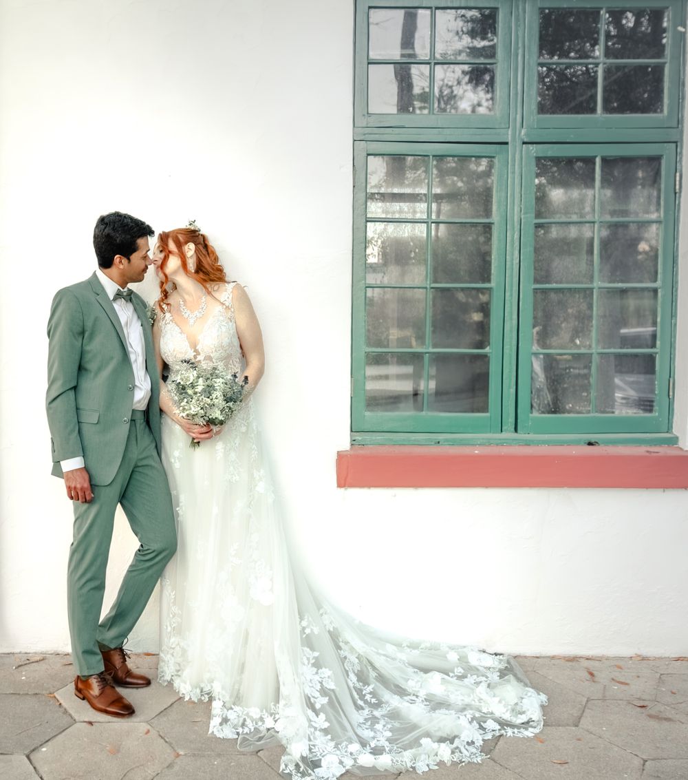 A couple in wedding attire poses together against a white wall with a mint green window frame.