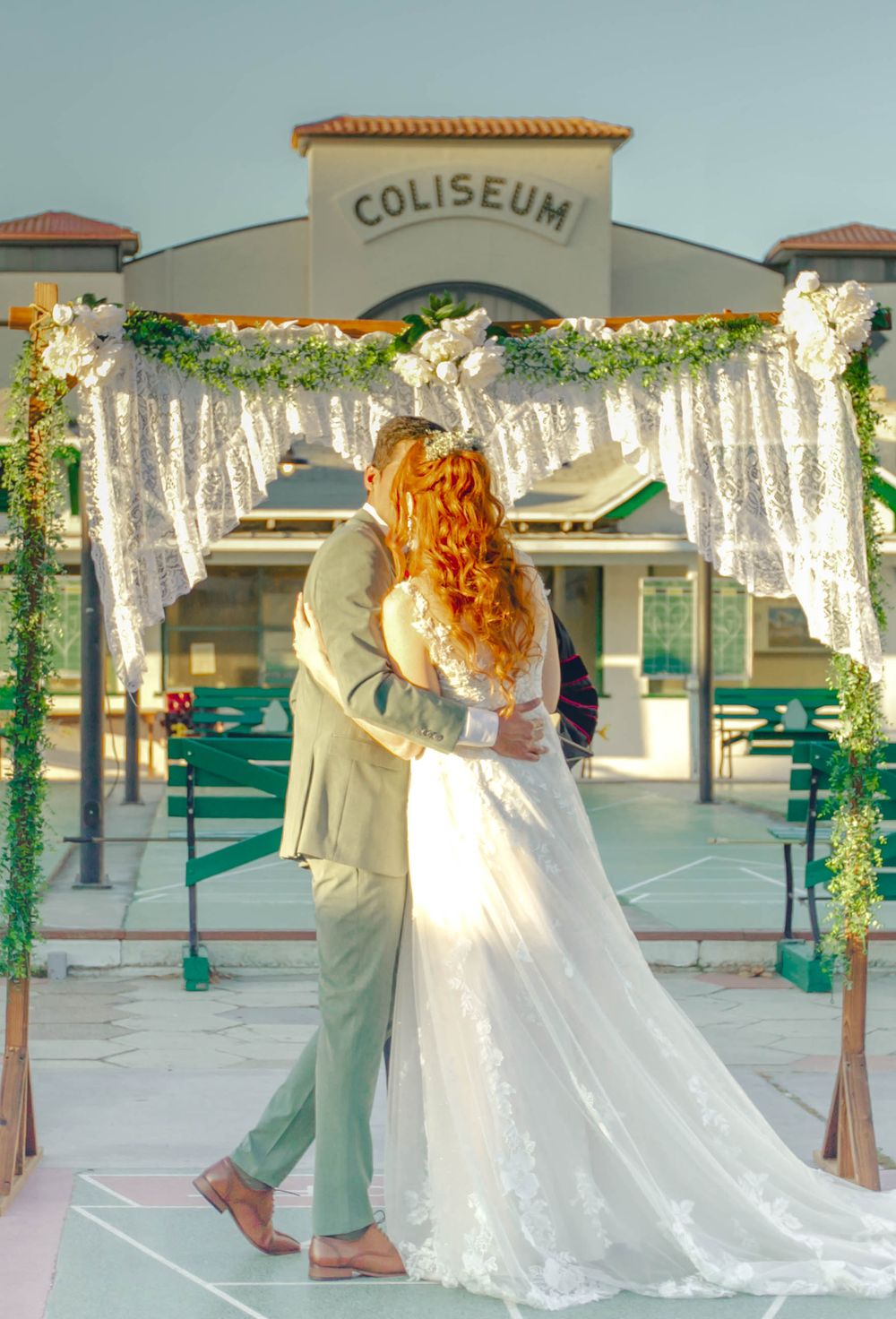 A bride and groom share a romantic moment under an ornate floral arch at the Coliseum during their outdoor wedding ceremony.
