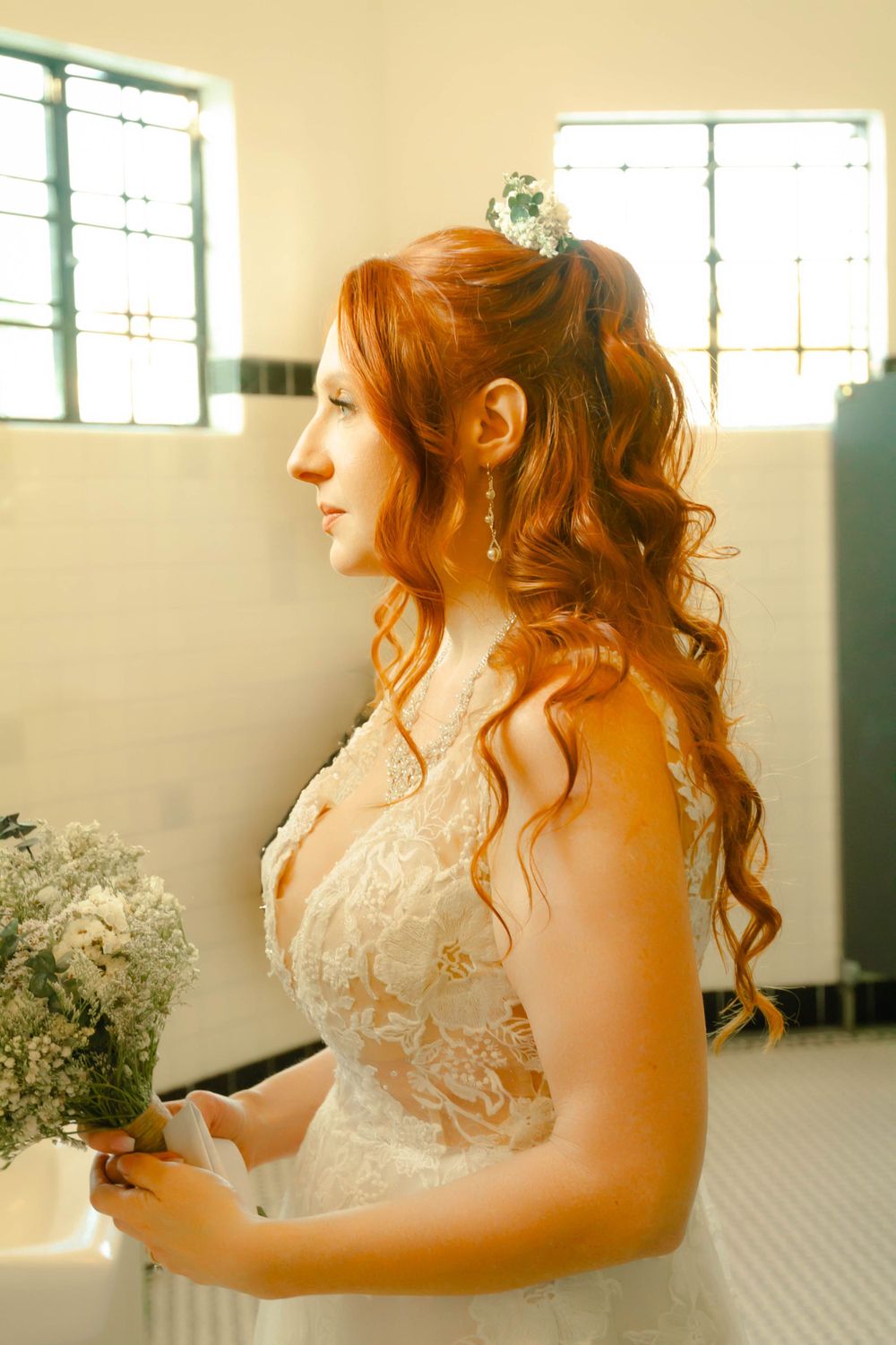 A bride in a lace wedding dress with long flowing curled hair styled in a romantic updo while holding dried flowers.