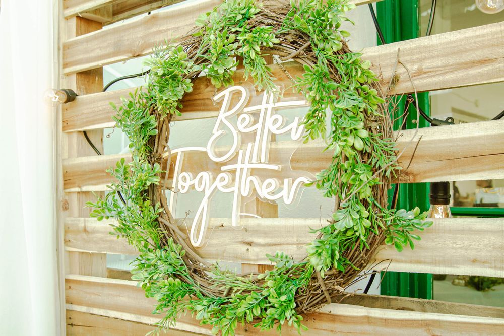 A decorative green wreath with neon 'Better Together' text sign mounted on a wooden pallet wall.