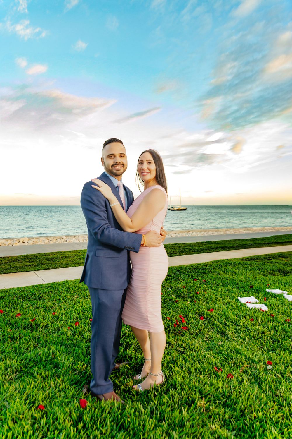 A couple embraces on a grassy lawn overlooking the ocean during sunset with a boat visible in the background.