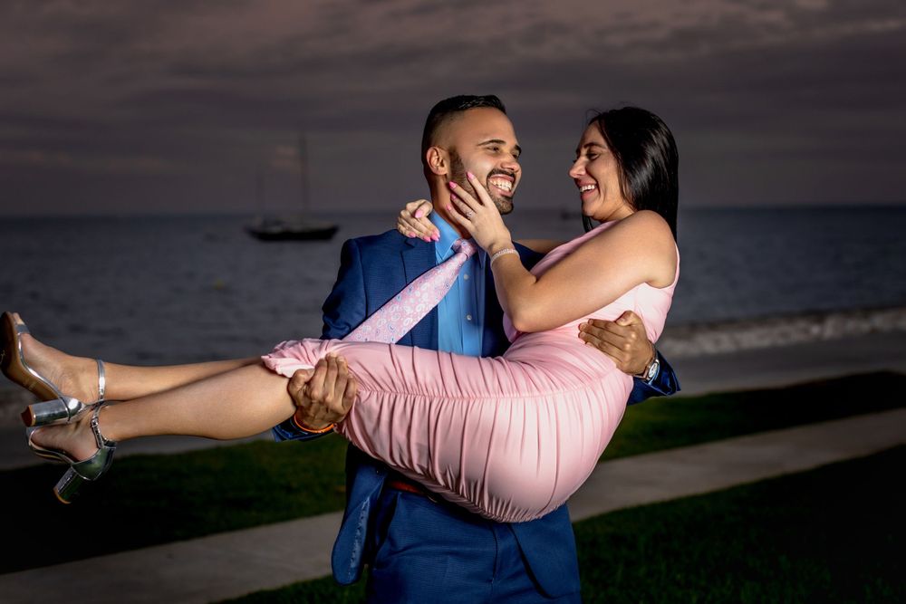 A romantic couple embraces in a bridal carry pose at sunset by the beach with a boat in the background.