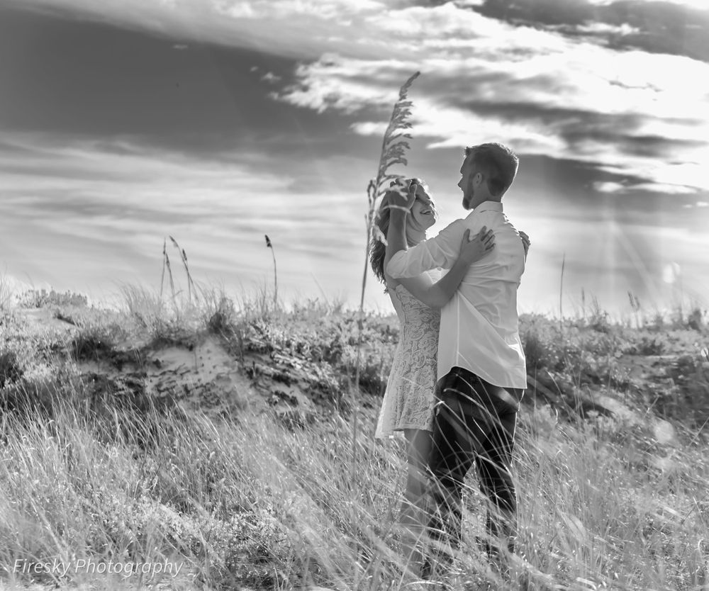 Black and white photo of couple standing in tall grass with dramatic sky.
