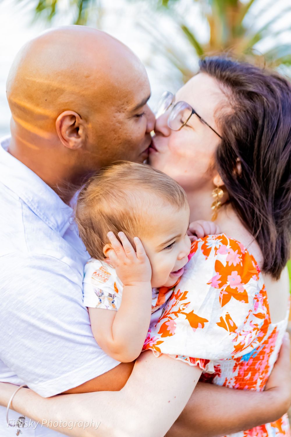 An intimate family moment as parents kiss while holding their smiling baby.