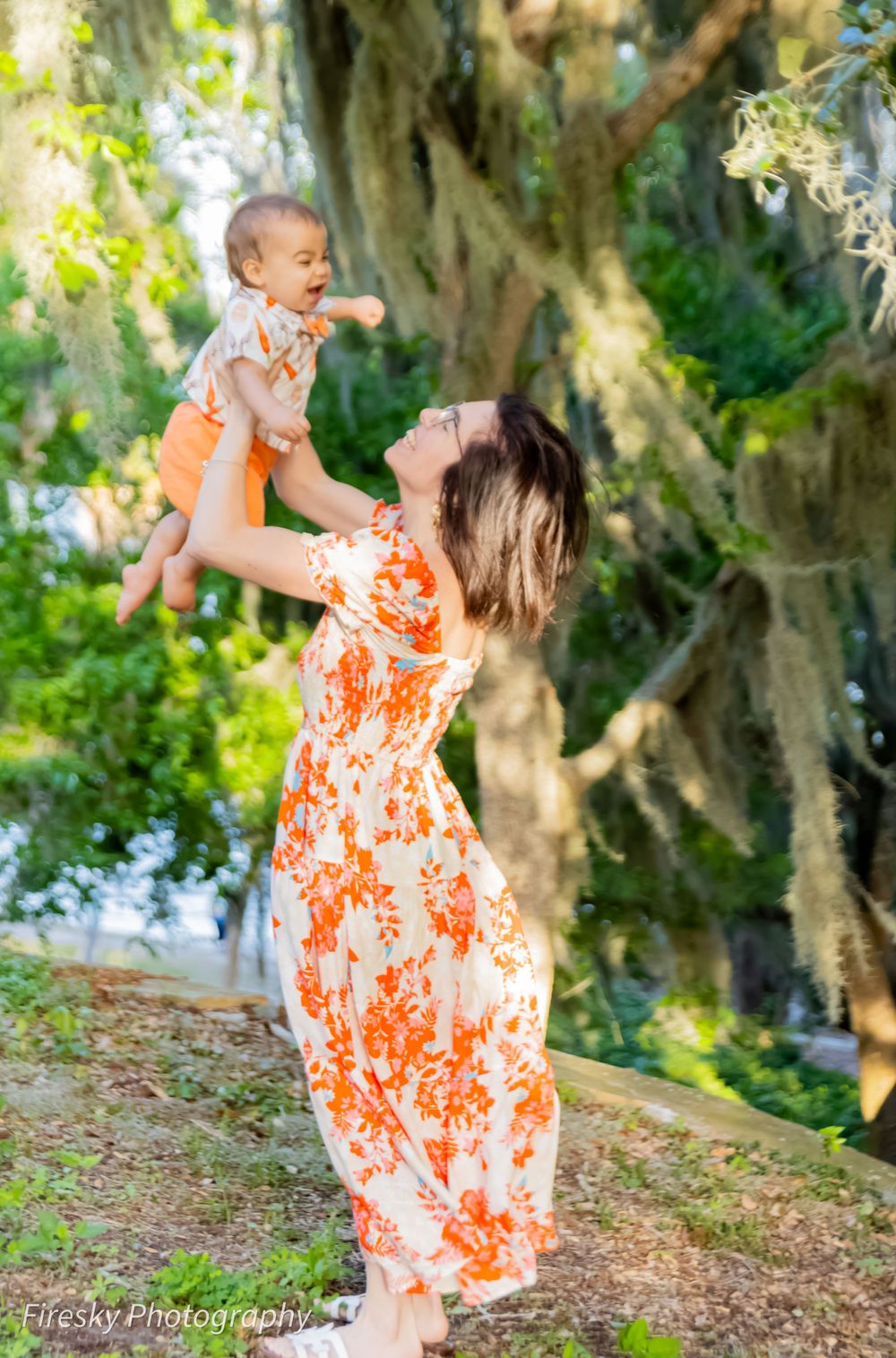 A joyful moment in a garden as someone wearing an orange floral dress lifts a baby up against a backdrop of Spanish moss.