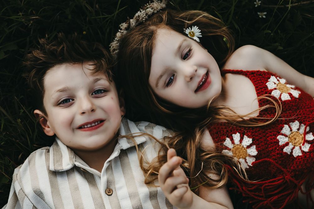 Two young children lying down together outdoors with one wearing a white shirt and one in a floral red dress.