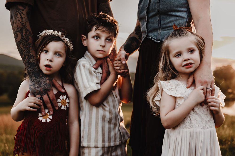 Three children in vintage-style formal attire stand together outdoors at golden hour.