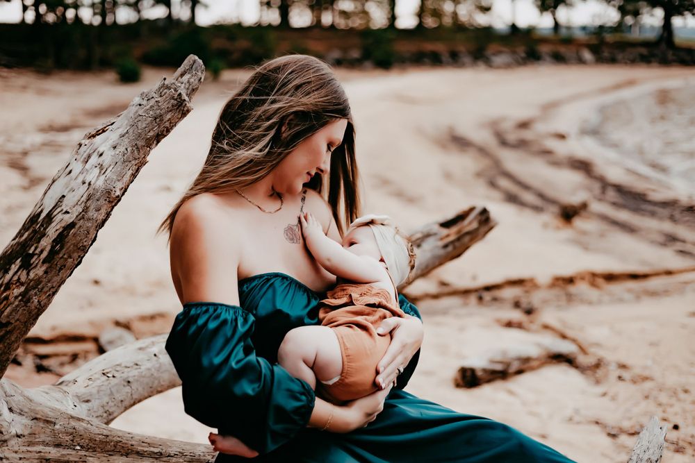 Parent and baby share a tender nursing moment on driftwood at a sandy beach in elegant teal dress.
