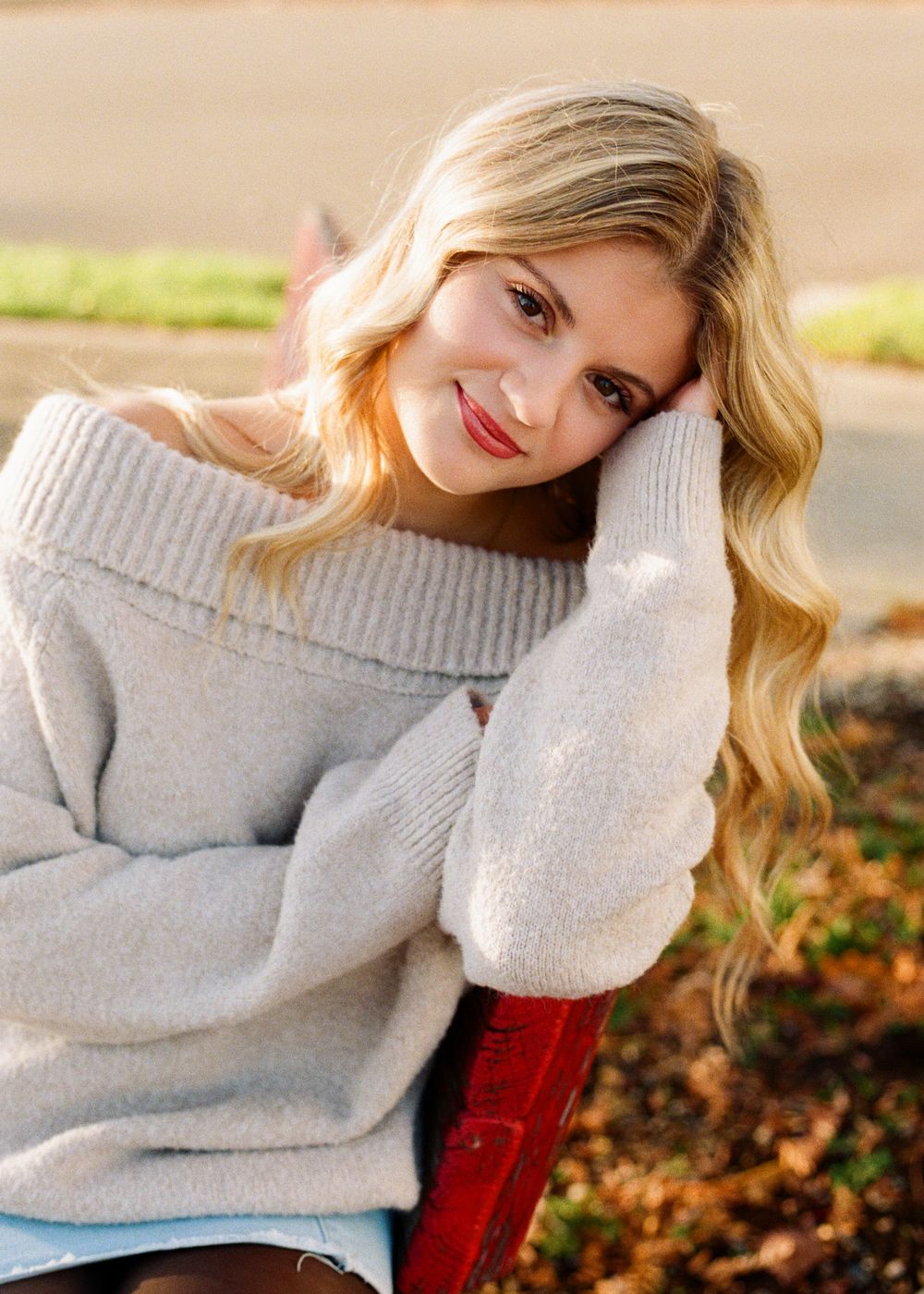 Person in cozy gray sweater poses outdoors on wooden bench with autumn leaves in background.