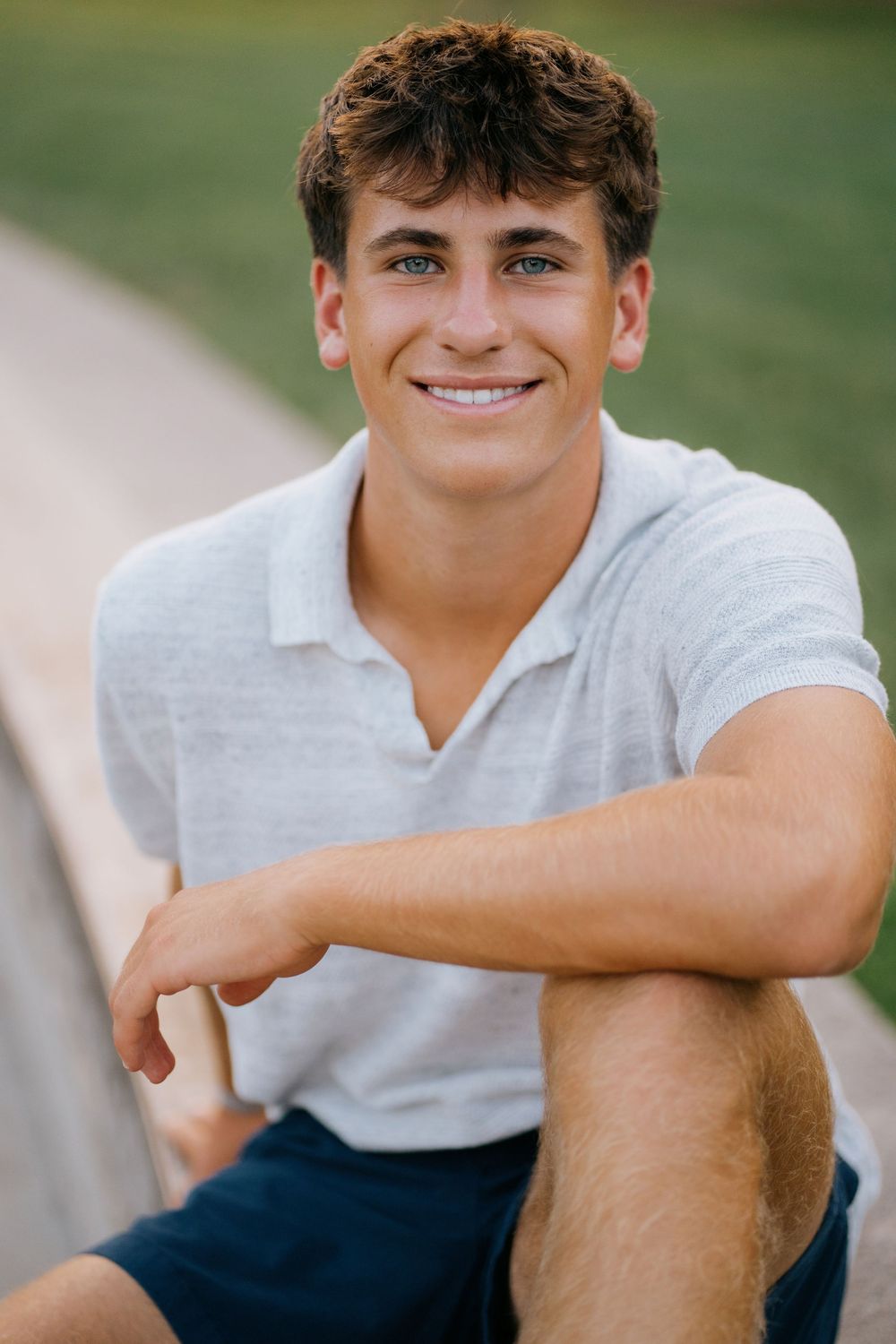 A person in a white polo shirt and dark pants poses casually outdoors against a blurred green background.