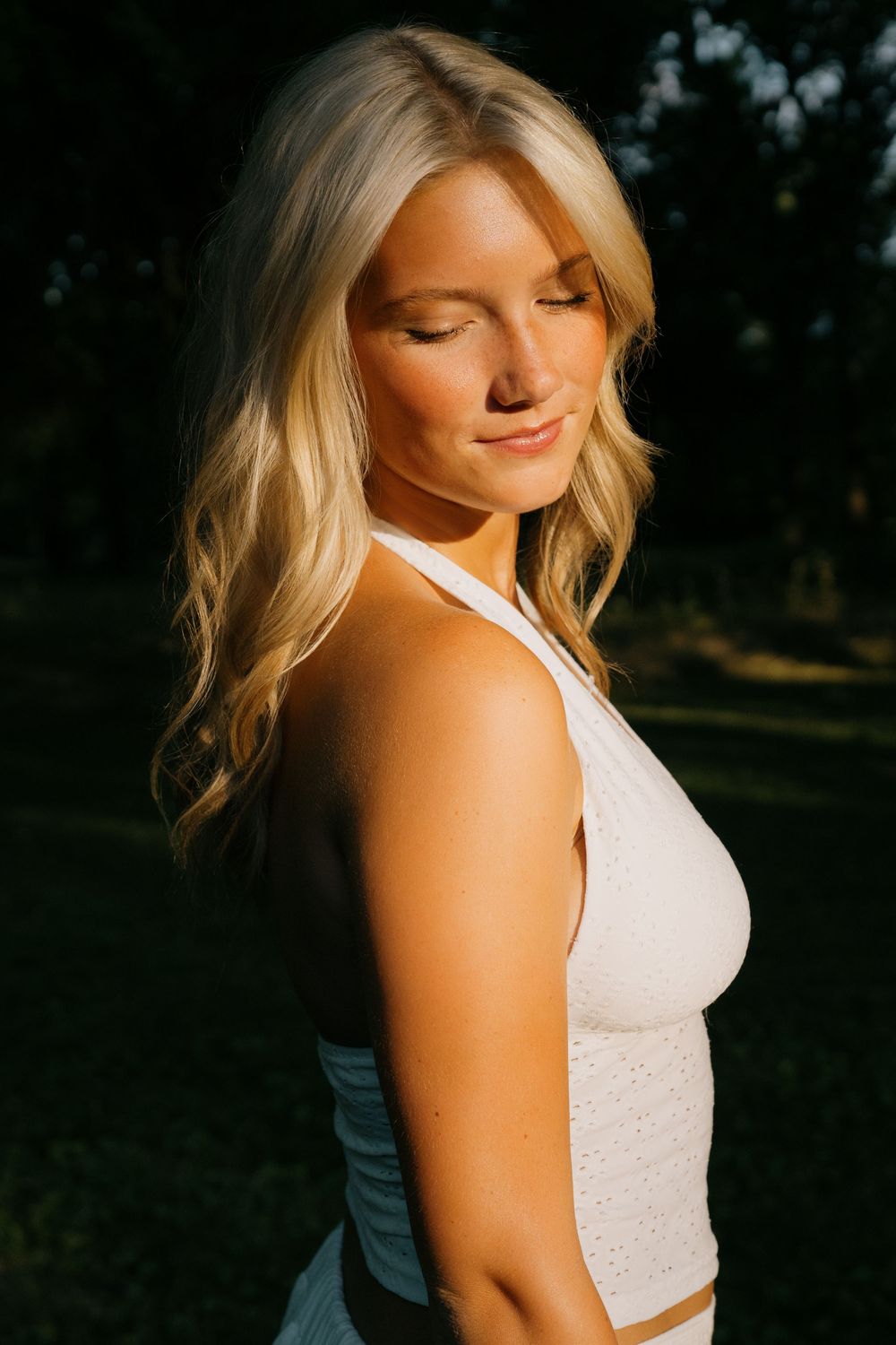 Blonde woman in white top enjoying golden hour sunlight outdoors, eyes closed, with lush green trees in background.