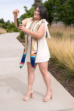 Graduate in white dress and serape stole celebrates with champagne bottle outdoors.