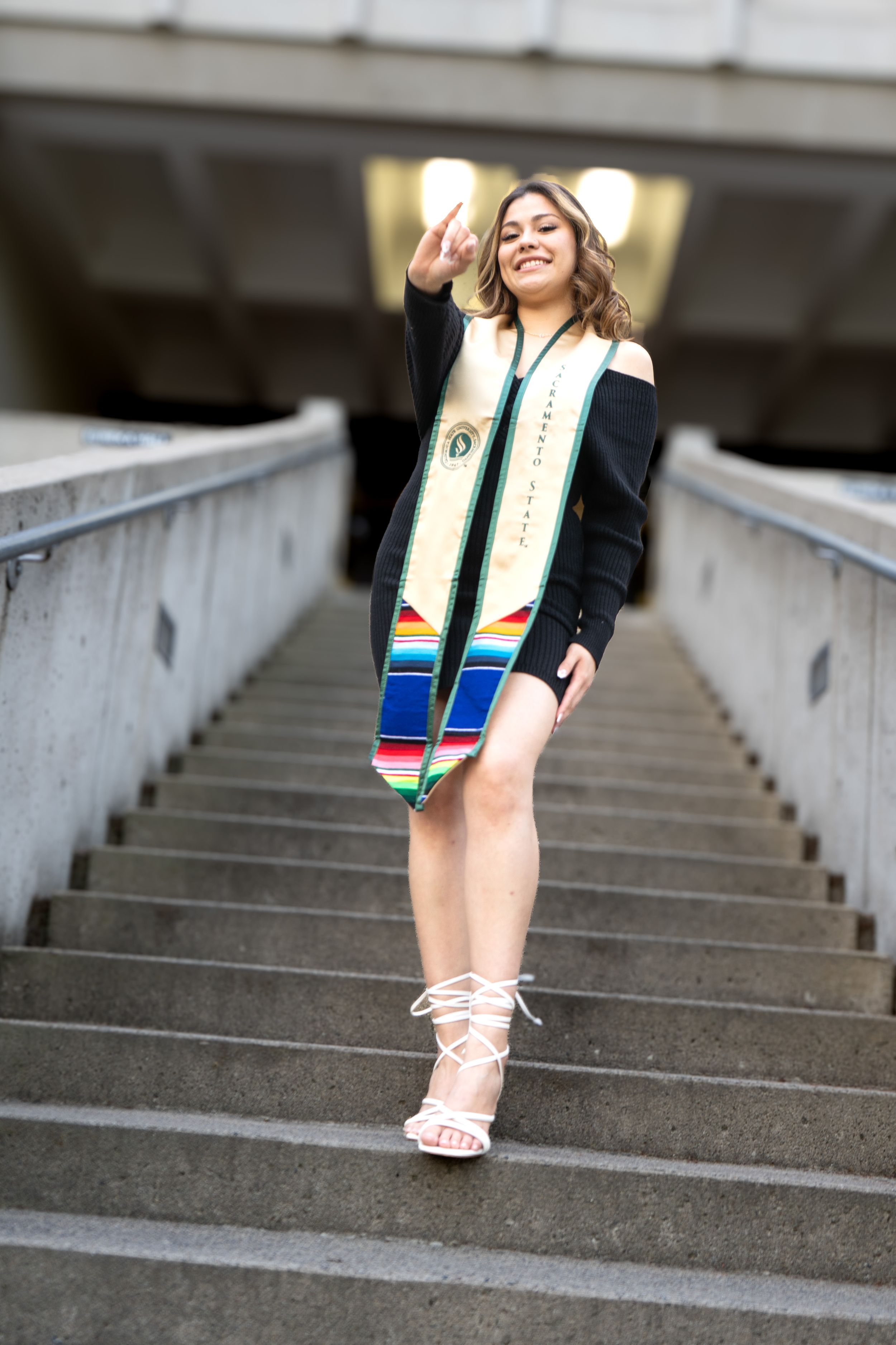 Young woman in graduation stole and black dress poses confidently on concrete steps.