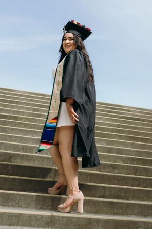 Graduate in black cap and gown with colorful stole poses on outdoor stairs.