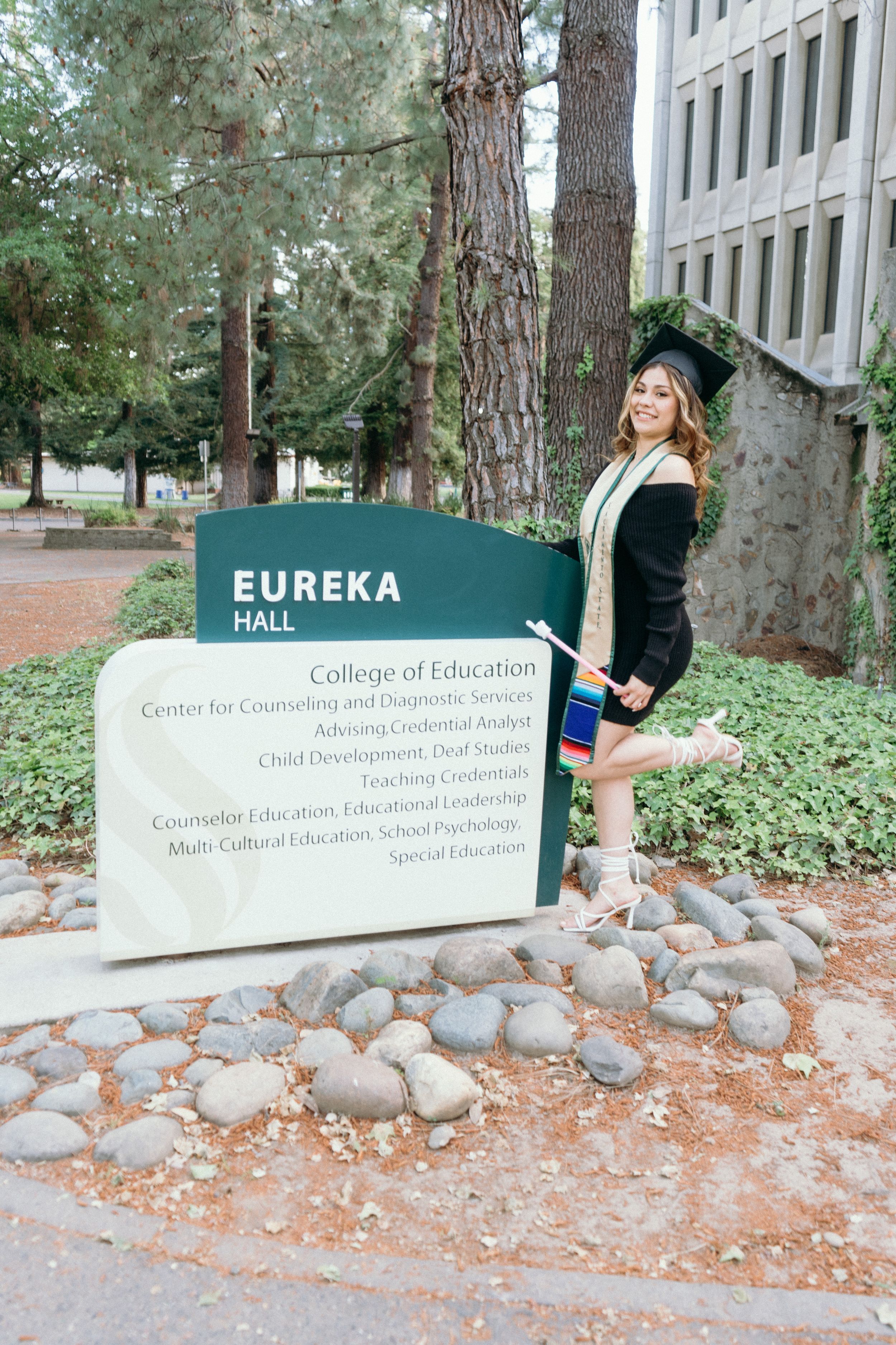 Proud graduate poses beside Eureka Hall College of Education sign on campus.