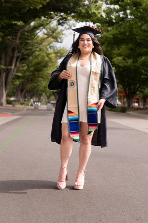 Graduate in black cap and gown with serape stole walks on campus pathway.