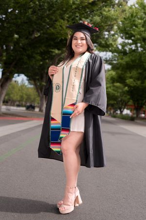 Smiling graduate in black cap and gown displays colorful serape stole on campus.