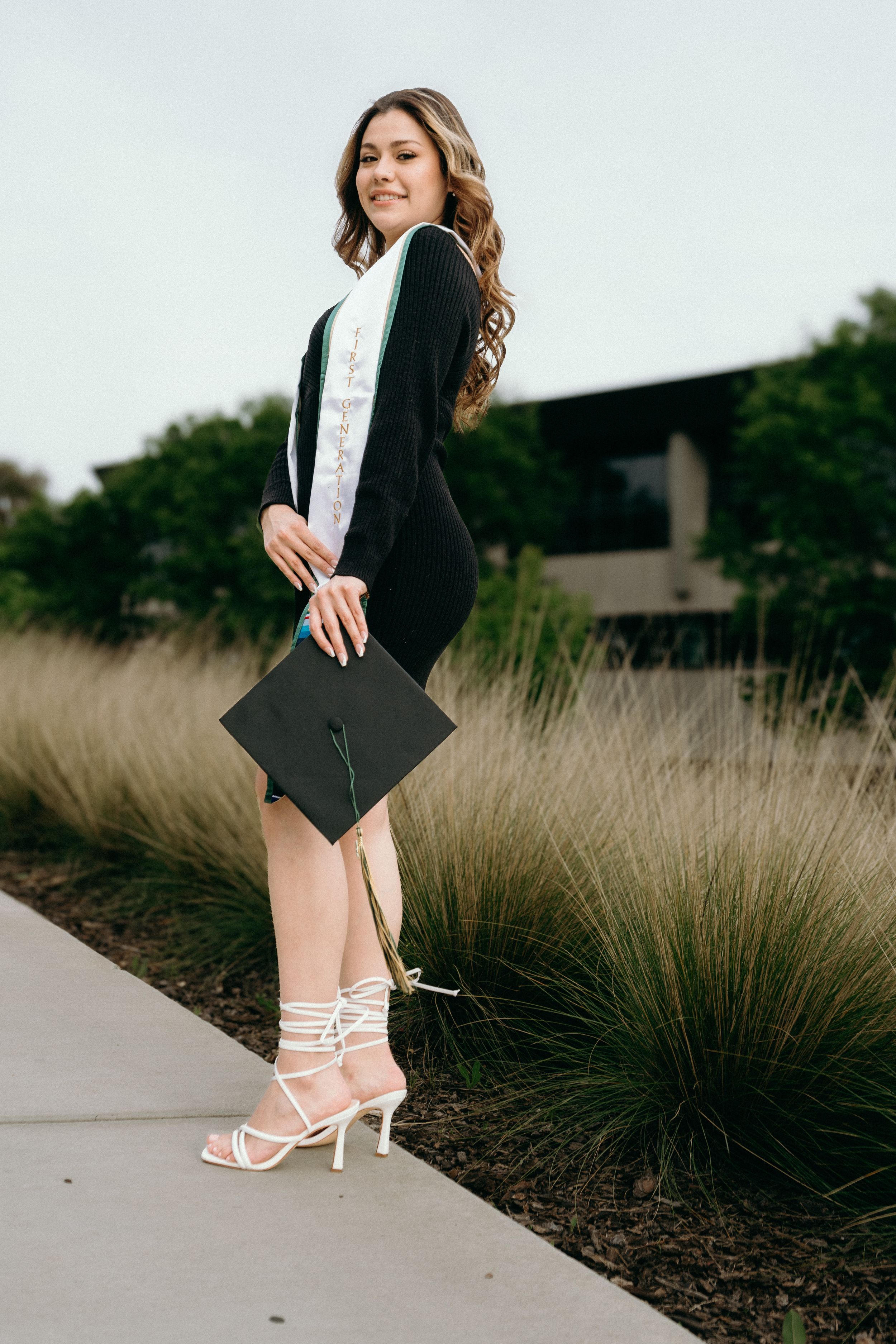 Female graduate in black gown holds mortarboard cap while posing on campus walkway in white heels.