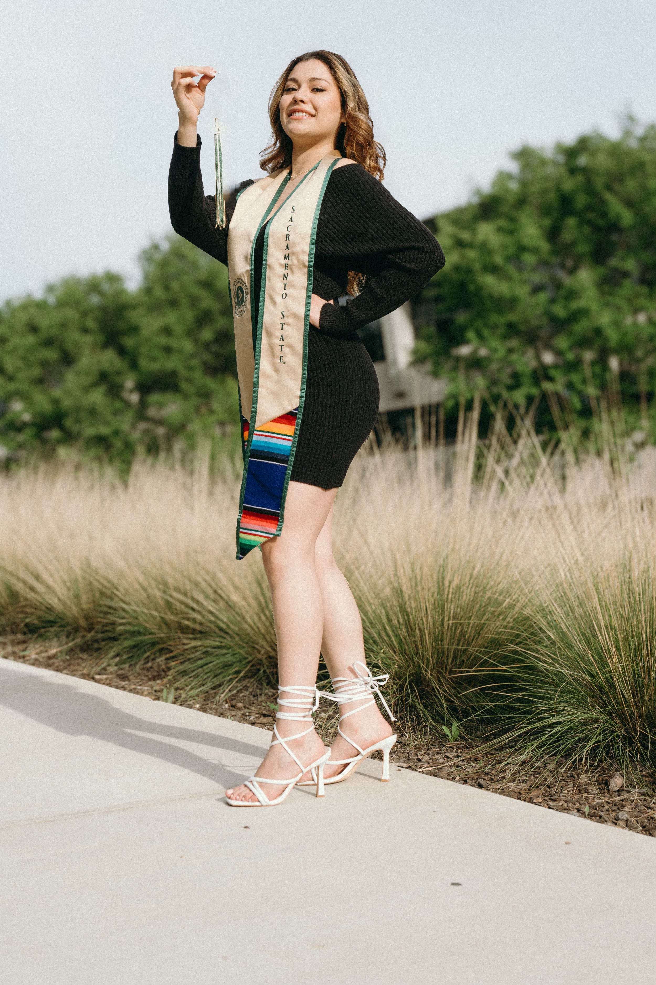 Smiling graduate in black dress holds tassel wearing colorful serape graduation stole on campus path.