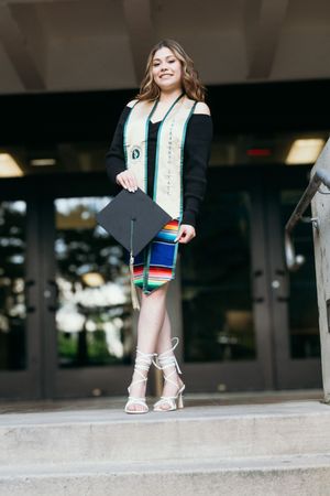 Graduate in black dress holds cap while standing on steps outside building entrance.