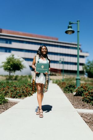 Smiling graduate in white dress holds green diploma cover while walking on sunny campus pathway.