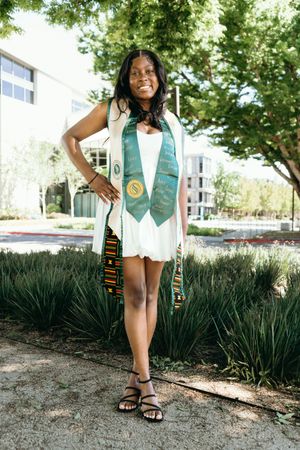 Smiling graduate in white dress with green and kente graduation stoles poses outdoors on campus.