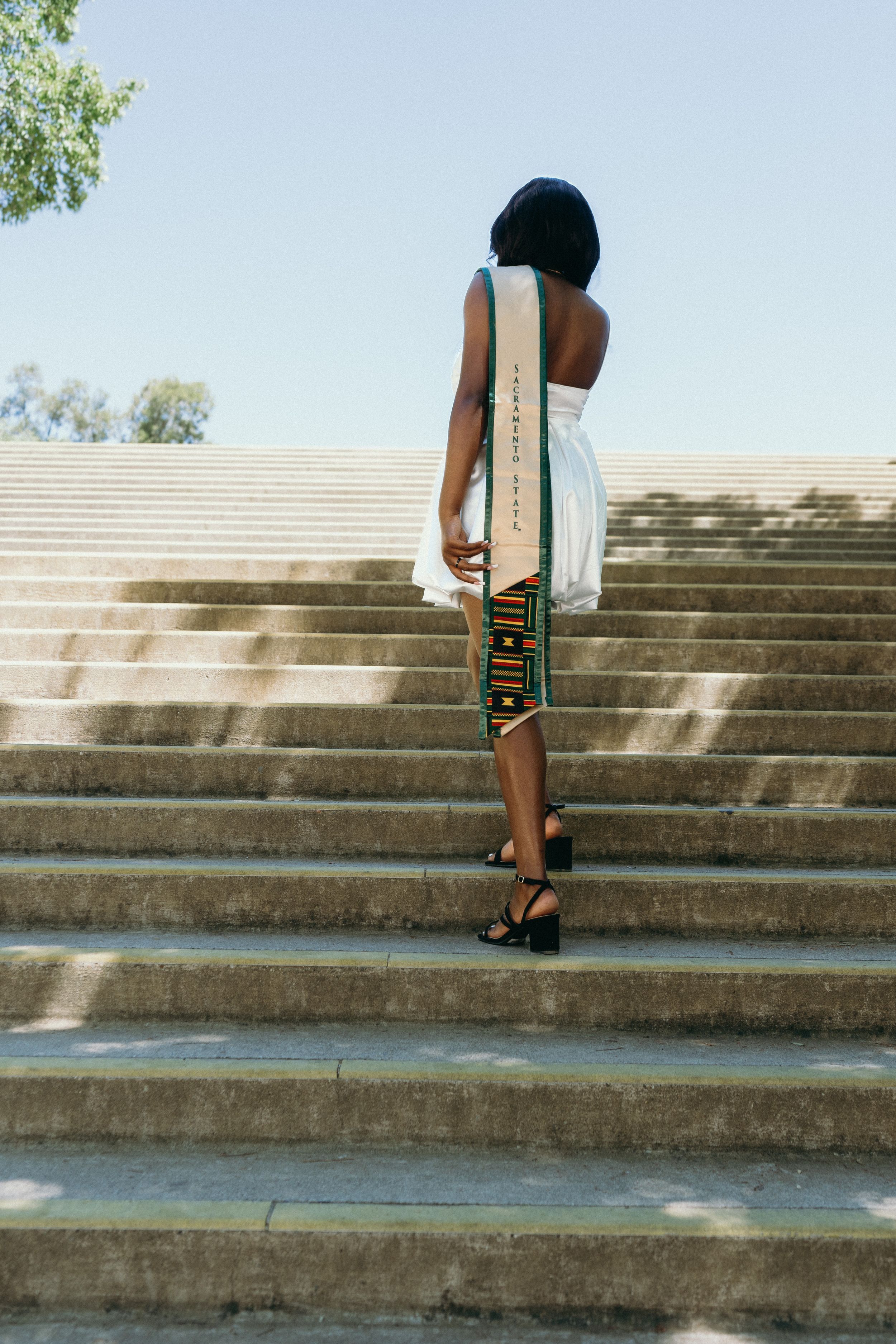 Graduate in white dress and kente stole stands with back to camera on stone steps.