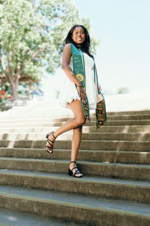 Young Black woman in white dress with green graduation stole poses on outdoor steps.