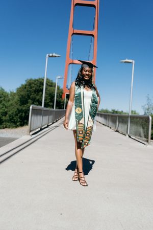 Graduate in white dress and cap walking on bridge with red structure against clear blue sky.