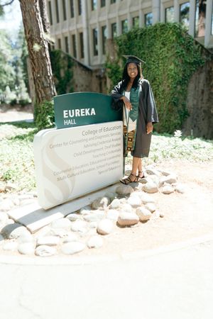 Graduate poses in cap and gown beside Eureka Hall College of Education sign at Cal State Sacramento.